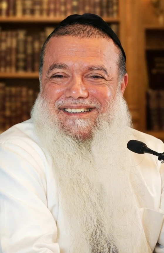 הרב יגאל כהן מחייך, בחולצה לבנה, על רקע ארון ספרים